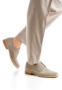 Chaussures en daim gris clair avec bouts arrondis et lacets ton sur ton. Une semelle en cuir marron présente un accent rouge. Portées avec un pantalon beige rayé.