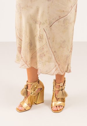 Sandalias de tacón alto doradas con correas adornadas, que cuentan con borlas y patrones intrincados, en contraste con un vestido floral claro.