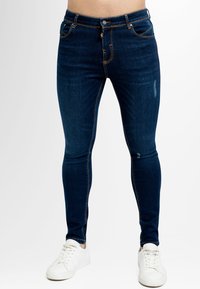 Jeans skinny azul oscuro de denim, con un clásico diseño de cinco bolsillos, costuras contrastantes y un cierre de botón, combinados con zapatillas blancas.