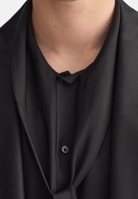 Camicia nera in tessuto setoso con scollatura drappeggiata e dettagli con bottoni. Texture liscia, caratterizzata da morbide pieghe e un'accatto a nodo sul colletto.