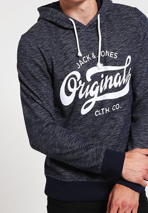 Marineblå hættetrøje med hvide snore og "Jack & Jones Originals CLTH. CO."-logo båret af en person med krydsede arme.
