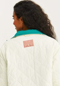 Prešívaná bunda v off-white farbe s tyrkysovým golierom, na zadnej strane s obdĺžnikovou oranžovou nálepkou s textom. Mäkká textúra a diamantový vzor.