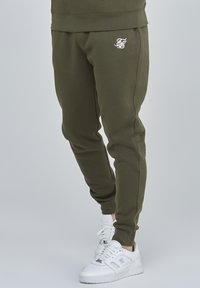 Olivegrüne Sweatpants aus weichem Stoff mit einem gestickten Logo am linken Oberschenkel und zulaufenden Beinen mit elastischen Bündchen.