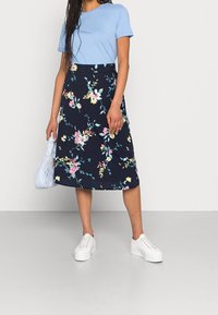 Jupe midi florale navy avec des fleurs roses et jaunes, associée à un t-shirt bleu clair, portée avec des baskets blanches et un sac à main bleu clair.