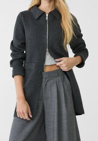 Dunkelgraue Jacke mit Reißverschluss, Kragen, gerippter Textur, langen Ärmeln und Seitentaschen, die über einem hellgrauen Oberteil und grauen Hosen getragen wird.