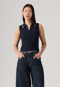 Haut polo sans manches en maille bleu marine avec col et ourlet blancs contrastants, associé à un jean en denim foncé à jambes larges présentant des détails de couture visibles.