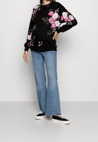 Persona vistiendo un sudadera negra con estampado floral rosa, jeans acampanados de color azul claro y zapatillas negras con suelas y cordones blancos.