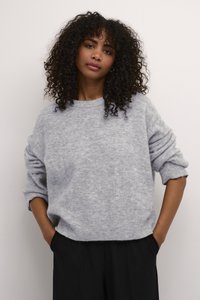 Lichtgrijze trui van zacht, textuurstof met een losse pasvorm, ronde halslijn en opgerolde mouwen. Gecombineerd met donkere broek, handen in de zakken.