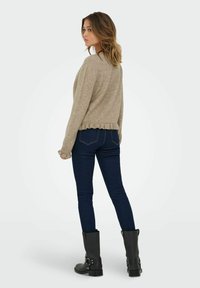 Bežs adīts džemperis ar apaļu kakla izgriezumu un ribotu apakšmalu, saskaņots ar tumši ziliem šaurajiem džinsiem un melniem potīšu zābakiem ar metāliskām detaļām.