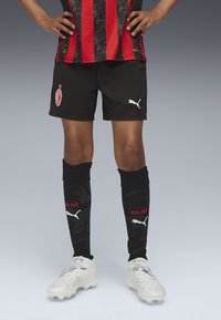 Schwarze Fußballshorts mit einem rot-schwarz gestreiften Trikot, akzentuiert mit einem Vereinslogo. Schwarze Socken mit einem Design und rotem "MILAN". Weiße Fußballschuhe.