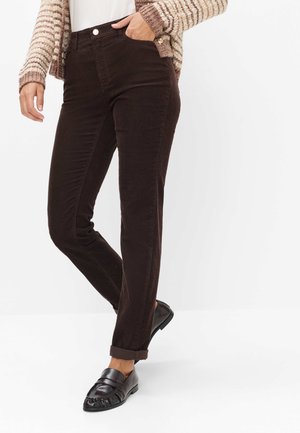 Femme portant un pantalon en velours côtelé marron foncé avec des revers, des mocassins noirs et un pull tricoté beige rayé, debout avec une main dans la poche.