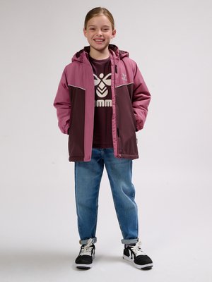 Bambino sorridente in piedi con giacca con cappuccio rosa e marrone, maglietta grafica bordeaux, jeans blu e scarpe da ginnastica nere e bianche su uno sfondo semplice.