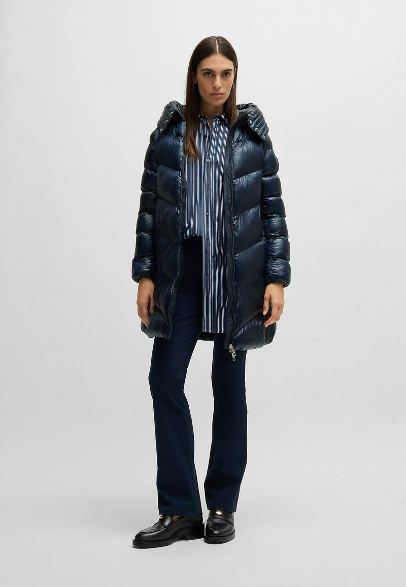 Granatowa kurtka puchowa o błyszczącym wykończeniu, z poziomymi przeszyciami i kapturem. Dobrana do niej jest koszula w paski, ciemne spodnie i czarne buty.