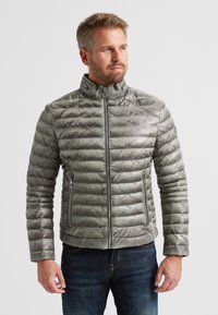 Grau gepolsterte Jacke mit hohem Kragen, Reißverschluss und Seitentaschen. Hat eine gesteppten Textur und glatte Oberfläche. Getragen über dunklen Jeans.