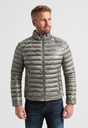 Grau gepolsterte Jacke mit hohem Kragen, Reißverschluss und Seitentaschen. Hat eine gesteppten Textur und glatte Oberfläche. Getragen über dunklen Jeans.