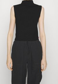 Schwarzes ärmelloses Oberteil mit hohem Ausschnitt, aus einem weichen, eng anliegenden Stoff gefertigt. Kombiniert mit dunklen, locker sitzenden Hosen mit Seitentaschen.