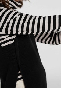 Zwarte mouwloze trui gelaagd over een zwart-wit gestreept shirt met lange mouwen. De materialen lijken gebreid met een gestructureerde afwerking.