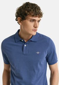Mörkblå polo skjorta i bomullsmix, med klassisk krage, två knappar och broderad logotyp på vänster bröst. Korta ärmar.