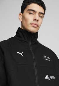 Sort zip-jakke med høj krave, der har hvidt Puma-logo og BMW Motorsport-patch. Materialet ser blødt ud med en glat tekstur.