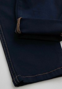 Granatowe spodnie z podwiniętymi mankietami, z kontrastującymi jasnobrązowymi szwami oraz gładką teksturą materiału. Brak widocznych elementów metalowych lub wzorów.