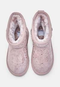 Ružové semišové členkové čižmy s umelou kožušinou, s malými okrúhlymi výrezmi a lesklými akcentmi na hornej časti. Okrúhla špička a prešité okraje.