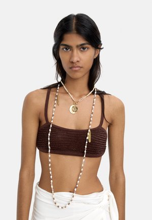 Młoda kobieta ubrana w brązową krótko dzianą bluzkę, nałożone warstwowo złote i muszelkowe naszyjniki oraz białą spódnicę z kopertowym zapięciem na tle jednolitego, jasnego tła.