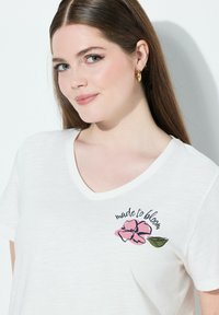 Camiseta blanca de manga corta con escote en V, que presenta un diseño floral en tonos rosas y el texto "hecha para florecer" en cursiva negra. Tejido suave y texturizado.