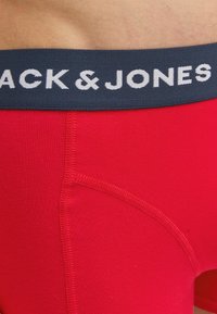 Rote Baumwollboxershorts mit einem marineblauen elastischen Bund, der die weiße Schrift "BLACK & JONES" trägt. Glatte Textur und figurbetontes Design.