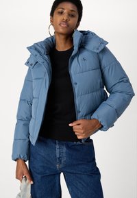 Giacca puffer azzurro chiaro con cappuccio removibile, chiusura con zip e tasche frontali. Presenta un motivo trapuntato e un dettaglio del logo sul petto.