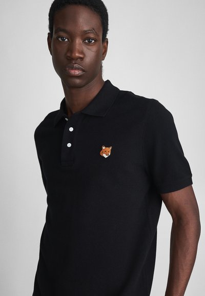 Un jeune homme à la peau foncée portant un polo noir avec une petite tête de renard orange brodée sur la poitrine.