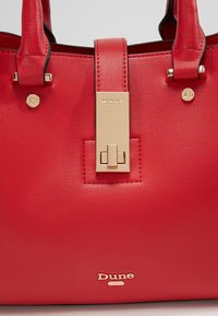 Sac à main en cuir rouge texturé avec fermoir couleur or, plaque logo et double anse. Marque Dune London visible au centre devant.