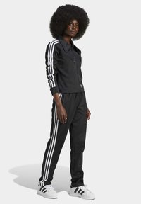 Fato de treino preto com riscas brancas nas mangas e nas laterais das calças. Apresenta uma jaqueta com fecho éclair, corte descontraído e sapatilhas brancas clássicas.