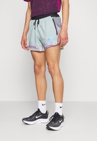 Pantalones cortos Nike Trail en azul claro con detalles morados, cinturilla elástica, cordón y bolsillo lateral, combinados con zapatillas deportivas negras.