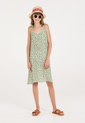 Groene bloemenjurk met dunne bandjes, knielengte, lichte stof, gecombineerd met een strohoed, ronde zonnebril en bruine sandalen.