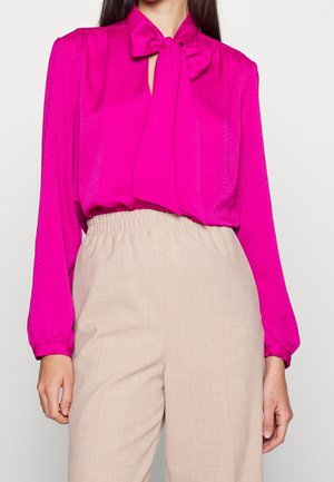 Donna che indossa una camicetta a maniche lunghe fucsia brillante con un fiocco annodato al collo e pantaloni beige a vita alta.