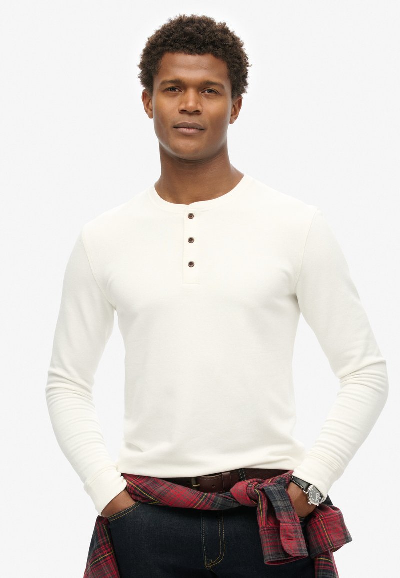 Cremfarbener Langarm-Henley-Shirt mit drei braunen Knöpfen, kombiniert mit dunkelblauer Jeans und einem rot karierten Hemd, das um die Taille gebunden ist.