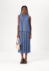 Rochie albastră plisată cu top fără mâneci și fustă fluidă. Pur și simplu cu sandale cu platformă negre și ținând o geantă mică bej.