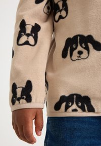Mano e avambraccio di un bambino che indossa una felpa in pile beige con stampi di facce di cani neri, abbinata a jeans di denim blu.