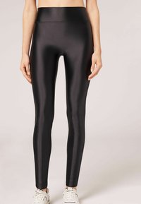 Leggings negros de cintura alta hechos de un material liso y brillante con un diseño ajustado, que se estrechan hasta los tobillos, combinados con zapatillas blancas.