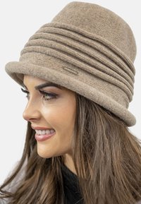 Beige Woll-Cloche-Hut mit strukturiertem Oberflächenmaterial, ausgestattet mit geschichteten Falten und einem kleinen metallischen Logo-Akzent. Glatter Rand, strukturierte Form.