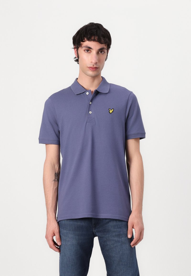 Violet polo marškinėliai su klasikine apykakle, trijų mygtukų užsegimu ir maža geltona siuvinėta logotipu ant krūtinės. Trumpomis rankovėmis.