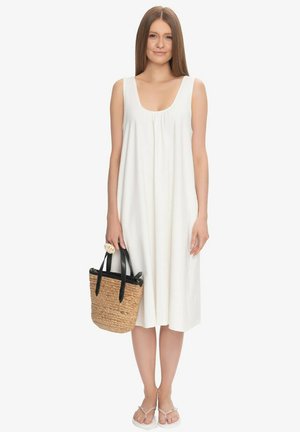 Kobieta ubrana w białą sukienkę midi bez rękawów, trzyma plecioną słomianą torbę z czarnymi uchwytami i nosi białe sandały.