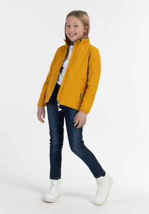 Giacca di pile gialla con colletto alto, chiusura a zip e tasche laterali, abbinata a jeans scuri e sneakers bianche alte.
