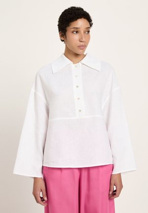 Woman with short curly hair wearing a loose white button-up shirt with wide sleeves and bright pink pants, looking to her left.