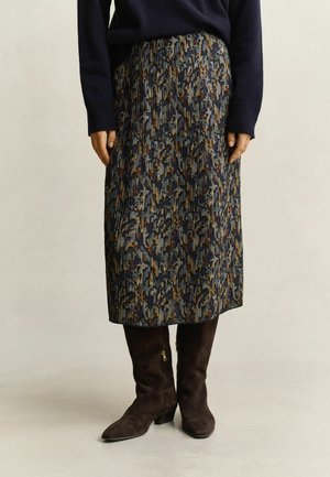 Femme portant une jupe midi plissée avec un motif abstrait multicolore, un pull sombre et des bottes en daim marron foncé mi-mollet avec petits talons.