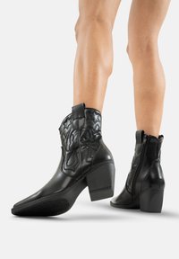 Botas de vaquero de cuero negro con punta afilada, patrón floral en relieve en el caña y tacón cuadrado. Cuenta con un zip lateral y una tira para facilitar su colocación.