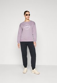 Light purple sweatshirt with white logo, paired with black sweatpants and beige sneakers. Model wears sunglasses, standing against a neutral backdrop.