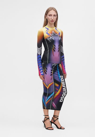 Femme portant une robe colorée à manches longues avec des motifs abstraits et le texte "DSQUARED2", associée à des sandales noires à talons hauts avec des lanières.