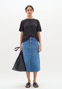 Svart kortärmad skjorta, blå denimmaxikjol, svarta slidesandaler och en stor svart läderväska, mot en enkel vit bakgrund.