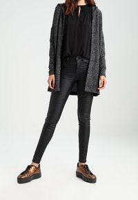 Černá blůza, dlouhý šedý cardigan, černé kožené kalhoty s ribbed detaily a hnědé platformové tenisky s černými tkaničkami, umístěné na neutrálním pozadí.